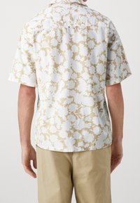 Camicia a maniche corte in beige chiaro con stampa floreale bianca. Presenta un colletto e una vestibilità rilassata, realizzata in tessuto traspirante con una finitura texturizzata.