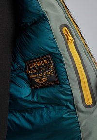 Interno della giacca imbottita con fodera teal, tessuto grigio, zip gialla e un'etichetta nera con testo arancione che indica marca e dettagli del prodotto.