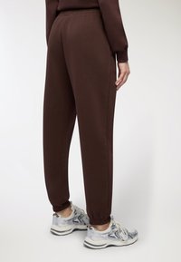 Brązowe dresy z elastycznym pasem i ściągaczami na dole, w połączeniu z jasnoszarymi i srebrnymi butami sportowymi z wzorzystym designem.