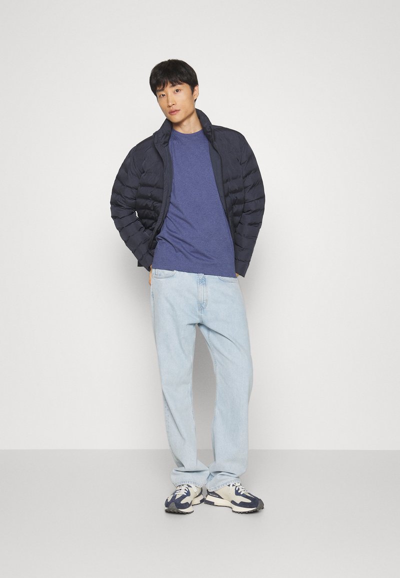 Marineblaue Puffjacke mit Stehkragen, kombiniert mit einem blauen T-Shirt, hellblauen Jeans und dunklen Sneakers mit weißem Akzent.