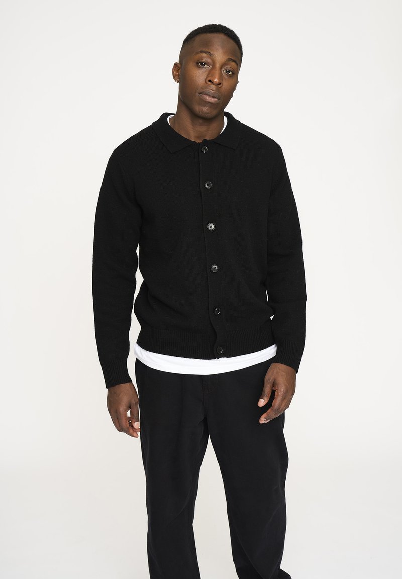 Sort cardigan lavet af tekstureret strikket stof, med krave, knaplukning fortil og ribbede manchetter, parret med en hvid T-shirt og sorte bukser.
