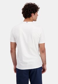 T-shirt bianca a maniche corte con colletto rotondo, caratterizzata da un piccolo accento rosso, bianco e blu sul colletto posteriore. Tessuto con superficie liscia.