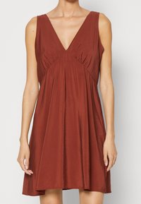 Robe couleur rouille en tissu léger avec un décolleté plongeant en V, sans manches, détails froncés sur les côtés, et une silhouette évasée en A.