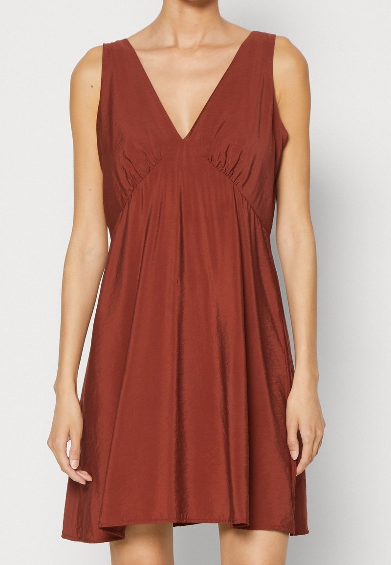 Robe couleur rouille en tissu léger avec un décolleté plongeant en V, sans manches, détails froncés sur les côtés, et une silhouette évasée en A.