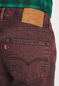 Vista trasera de unos jeans Levi's 501 rojos con parche de cuero de la marca, costuras visibles y etiqueta roja de Levi's debajo de una camisa de cuadro verde.