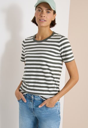 Femme portant un t-shirt rayé vert et blanc, un jean bleu clair et une casquette vert pâle, debout avec les mains dans les poches.