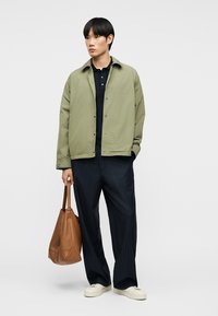Homme portant une veste vert clair sur une chemise noire, pantalon noir à fines rayures, baskets blanches, tenant un grand sac fourre-tout en cuir marron, debout.