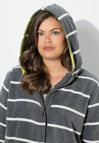 Felpa grigia con cappuccio in pile, con strisce bianche e una fodera interna giallo brillante. Presenta una chiusura con bottoni e cappuccio attaccato. Texture morbida.