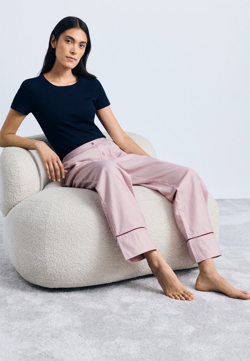 Marinblå T-shirt, rosa vida byxor med små fållaccent, sittande på en plyschig, beige skulptural stol mot en ljus bakgrund.