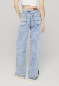 Persona valkā gaisā zilo plašu staru džinsus ar augstu jostasvietu, redzamām aizmugures kabatām un brūnu ādas zīmola plāksteri, kombinējot ar baltām sporta kurpēm.