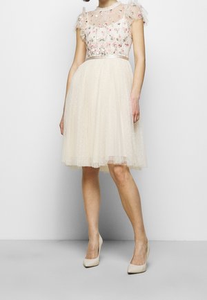 Femme portant une robe crème transparente longueur genoux avec broderie florale sur le corsage et manches courtes, associée à des chaussures à talons crème.