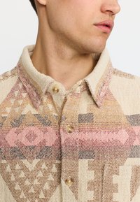 Beige gemustertes Hemd mit Kragen, das geometrische Designs in Rosa, Braun und Grau aufweist. Es hat hellfarbene Knöpfe und einen strukturierten Stoff.
