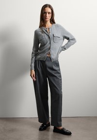 Gilet en tricot gris avec deux poches sur la poitrine, associé à un pantalon large gris foncé et des mocassins noirs sur un sol texturé.