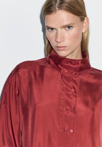 Red satin shirt with a high collar, front button closure, and smooth texture. Fabric has a slight sheen and a relaxed fit.