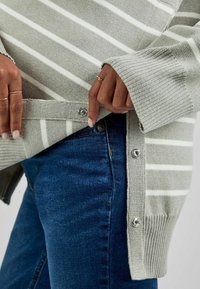 Grau-weiß gestreifter Pullover mit gerippten Bündchen, seitlichen Druckknöpfen und lockerer Passform, kombiniert mit dunkelblauen Jeans.