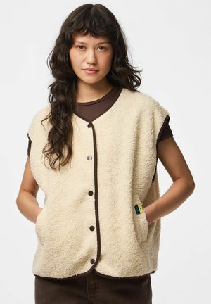 Femme portant un gilet sans manches en polaire beige avec des boutons-pression et une bordure marron sur un t-shirt marron à manches courtes, mains dans les poches du gilet.