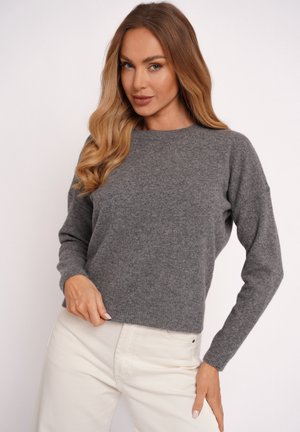 Woman with long blonde hair wearing a grey knit sweater and light beige pants, posing with one hand on hip and the other holding sweater hem.