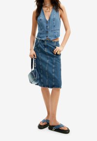 Jeansväst med djup V-ringning och knappstängning, i kombination med en knälång jeanskjol med vertikala sömmar. Mörkblå sandaler och en liten väska.