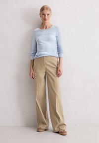 Lichtblauw langemouwen shirt met een vervaagde logo, gecombineerd met beige wijde broek en tan sneakers op een effen achtergrond.