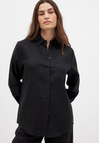 Black button-up shirt made of a textured fabric. Features a collar, long sleeves, and a small breast pocket. Simple design with a relaxed fit.