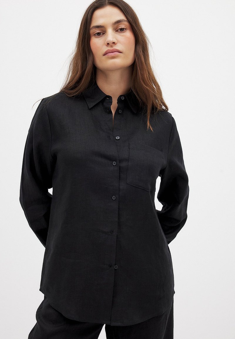 Black button-up shirt made of a textured fabric. Features a collar, long sleeves, and a small breast pocket. Simple design with a relaxed fit.