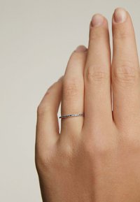 Anillo delgado de plata con una fila de pequeñas piedras azules, llevado en una mano de piel clara. Diseño simple, textura suave, estilo minimalista.