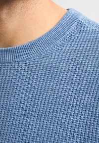 Pulover tricotat de culoare albastră deschisă, cu guler ribat, având un model texturat de tip waffle. Vedere de aproape care evidențiază detaliile țesăturii.