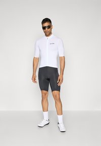 Maillot de cyclisme à manches courtes en blanc avec une fermeture éclair à l'avant, associé à un short de cyclisme noir et des chaussures blanches. Des lunettes de soleil accentuent le look sportif.