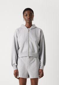 Felpa grigia con zip e cappuccio, tasche frontali e polsini a costine, abbinata a pantaloni corti grigi con vita elasticizzata.