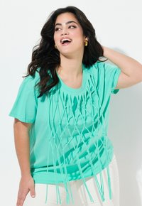 En lys turkis T-shirt har et knyttet design foran, med lange, frynsede detaljer der hænger ned. Korte ærmer og en rund halsudskæring.