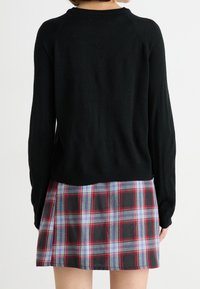 Personne portant un pull noir à manches longues et une jupe à carreaux rouge, bleu et blanc, vue de dos sur un fond blanc.
