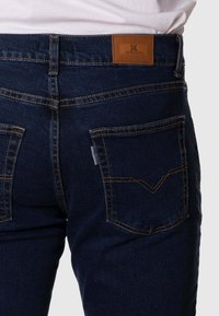 Vaqueros de mezclilla azul oscuro con una textura suave, que cuentan con dos bolsillos traseros y una etiqueta de marca de cuero marrón en la cintura. Costuras en contraste.