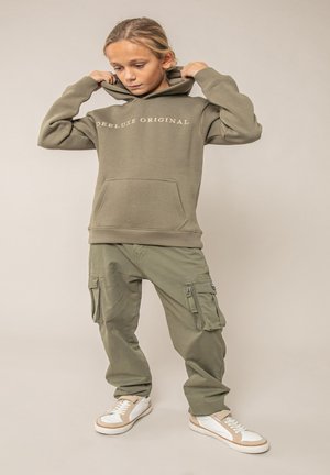 Enfant ajustant la capuche d'un sweat-shirt "Deluxe Original" vert olive, portant un pantalon cargo assorti et des baskets blanches sur un fond uni.