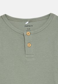 Ανοιχτό πράσινο ριγέ μπλουζάκι με στρογγυλή λαιμόκοψη, ξύλινη πατιλέτα με κουμπιά και ετικέτα που εμφανίζει πληροφορίες για το μέγεθος και τη μάρκα.