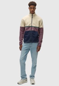 Chaqueta de forro polar con cuello alto, con paneles en crema, rosa y azul marino; cremallera completa; tejido texturizado; jeans de mezclilla azul claro; zapatillas beige.