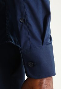 Ciemnoniebieski mankiet koszuli o gładkiej, matowej strukturze z dwoma czarnymi guzikami i przeszyciem wzdłuż krawędzi.