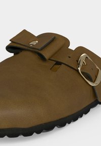 Sabot marron à enfiler avec une large lanière en cuir, une boucle dorée et une semelle noire texturée.