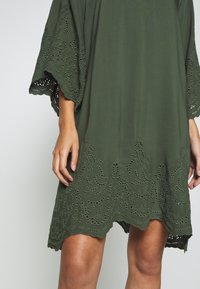 Olivgrünes Kleid mit aufwändigen Spitzenausschnitten am Saum und an den Ärmeln, mit lockerem Schnitt und fließendem Stoff.