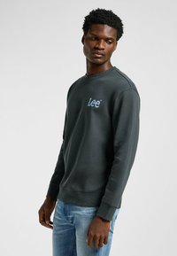 Felpa verde scuro con scollatura a girocollo a coste e polsini, caratterizzata da un logo ricamato blu "Lee" sul lato sinistro del petto. Indossata con jeans azzurri.