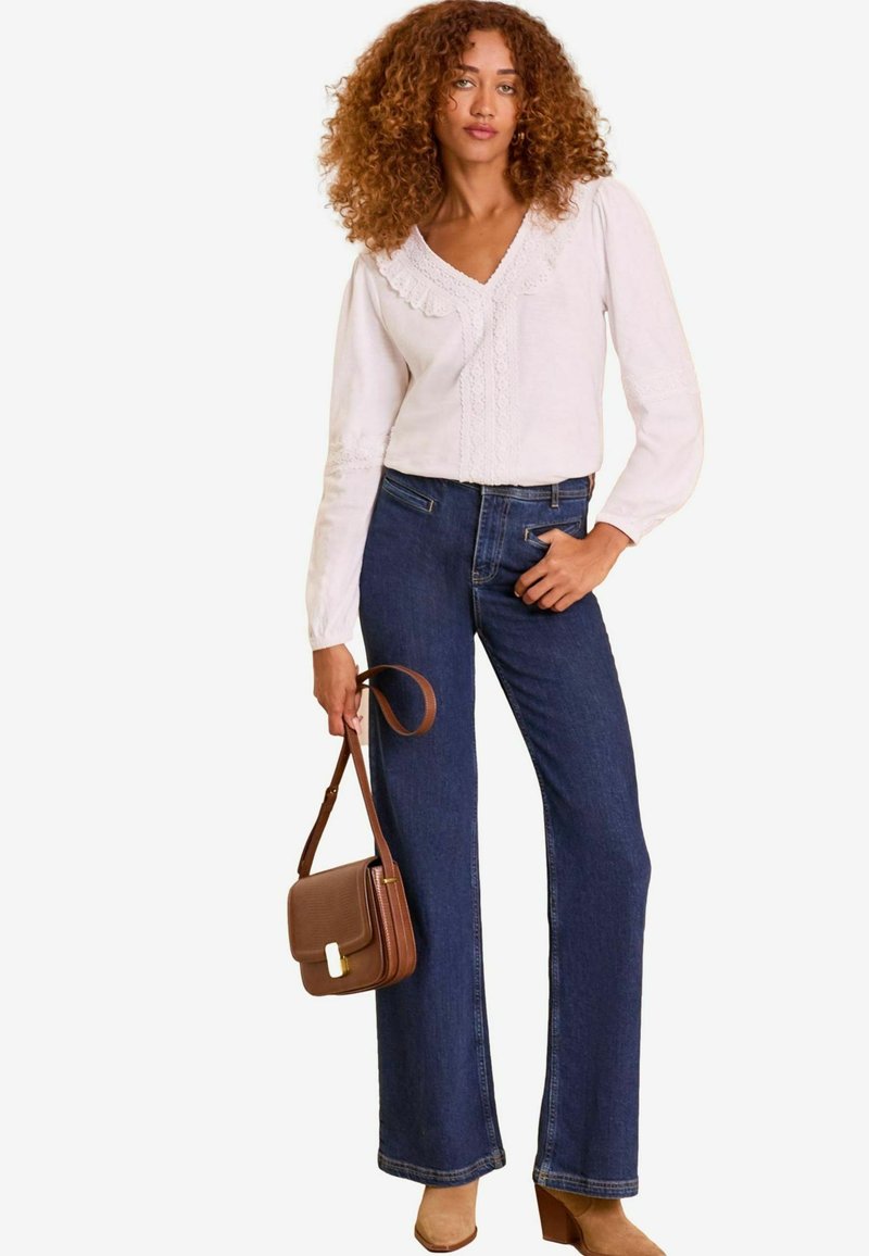 Jeans in denim blu a zampa d'elefante abbinati a una blusa bianca con dettagli in pizzo. Il modello tiene una borsa marrone e indossa stivali con tacco beige.