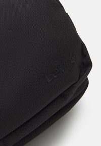 Primo piano di un angolo di tessuto nero testurizzato con il sottile logo ricamato Levi's su una borsa o un sacchetto morbido.