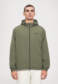 Oliven grøn zip-up jakke med hette, elastiske ermer og sidelommer; har et lite logo på brystet og glatt, lett stoff.
