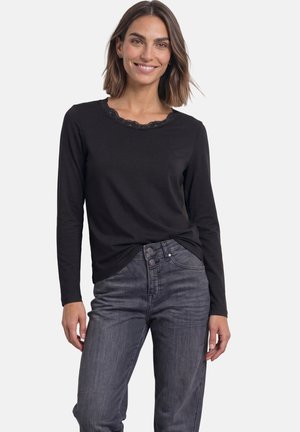 Kvinde med skulderlangt hår iført en sort langærmet top med blonder ved halsudskæringen og falmede grå jeans, smilende med afslappede hænder.
