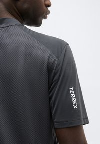 Άνδρας που φοράει σκούρο γκρι αθλητική κοντομάνικη μπλούζα με υφή στην πλάτη και την επιγραφή «TERREX» σε λευκό στο δεξί μανίκι.