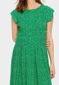 Vihreä mekko, jossa on lyhyet lepattelevat hihat, smockattu yläosa ja valkoisia pilkkuja. Hame on löyhästi ryhmitelty virtaavuutta varten.
