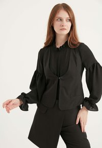 Schwarze Weste mit niedrigem V-Ausschnitt, Reißverschluss vorne und zwei Taschen, über einem schwarzen Oberteil mit gerafftem Kragen und Puffärmeln getragen.