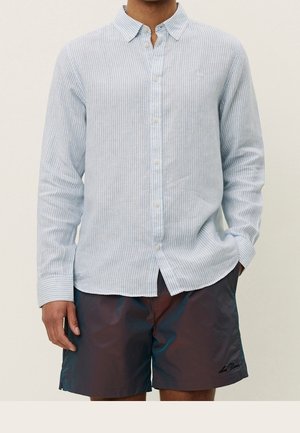 Homme portant une chemise à rayures bleu clair à manches longues boutonnée et un short foncé, les mains relâchées sur les côtés.