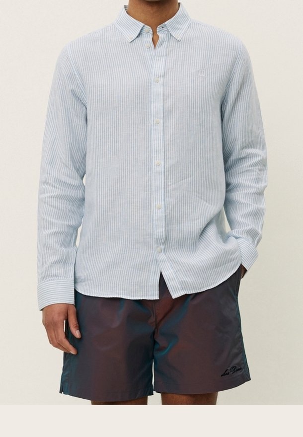 Homme portant une chemise à rayures bleu clair à manches longues boutonnée et un short foncé, les mains relâchées sur les côtés.