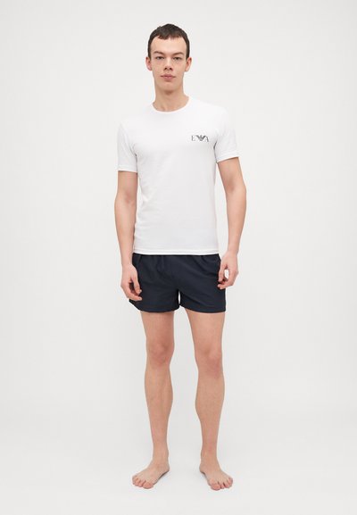 T-shirt blanche à manches courtes avec un logo noir sur la poitrine gauche, associée à un short marine foncé, portée par un modèle masculin debout, pieds nus.