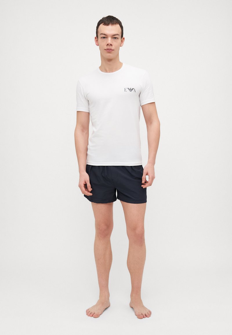 T-shirt blanche à manches courtes avec un logo noir sur la poitrine gauche, associée à un short marine foncé, portée par un modèle masculin debout, pieds nus.